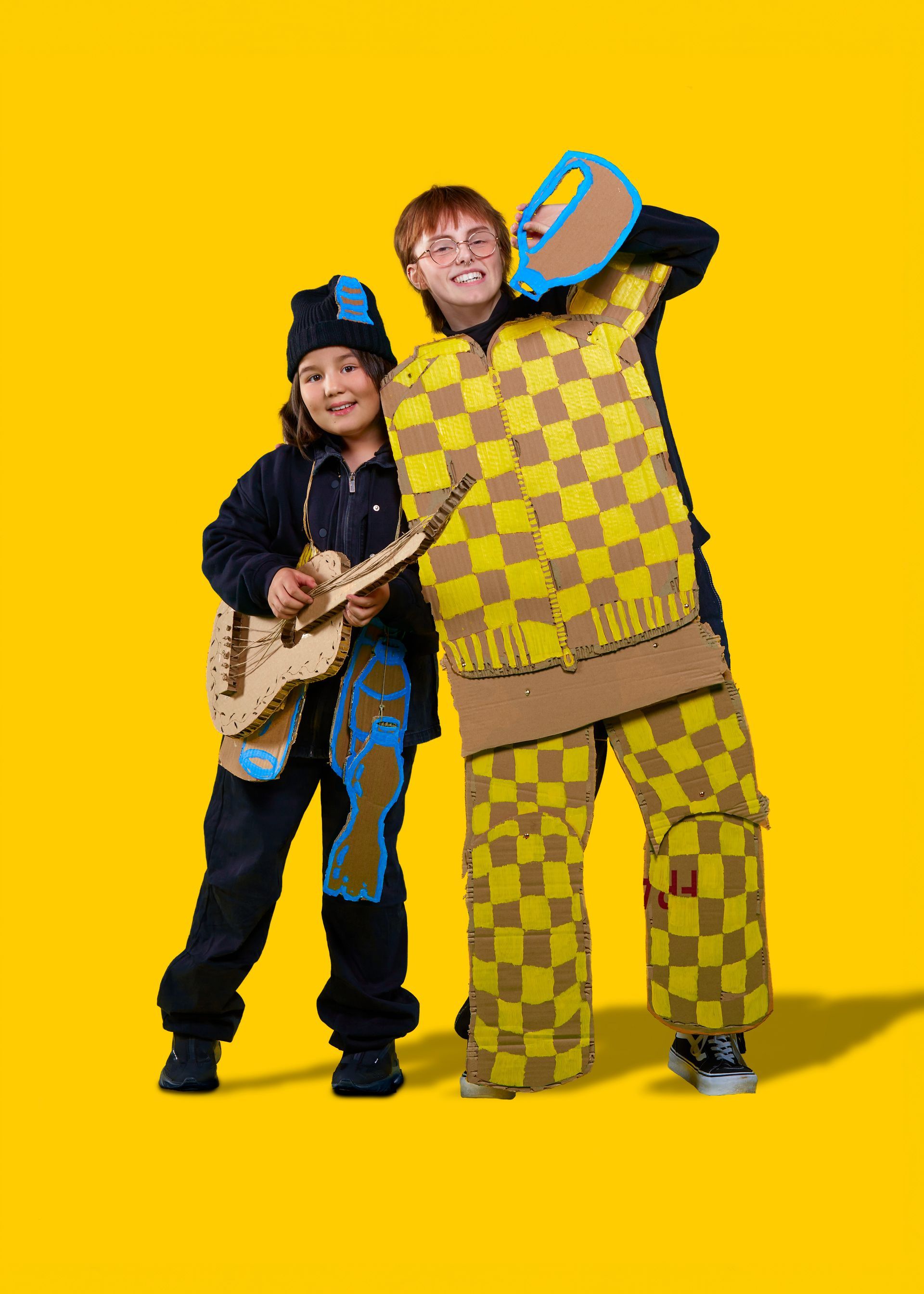Twee personen in geel-bruin geblokte outfits, waarvan één met een gitaar, lachend op een gele achtergrond.