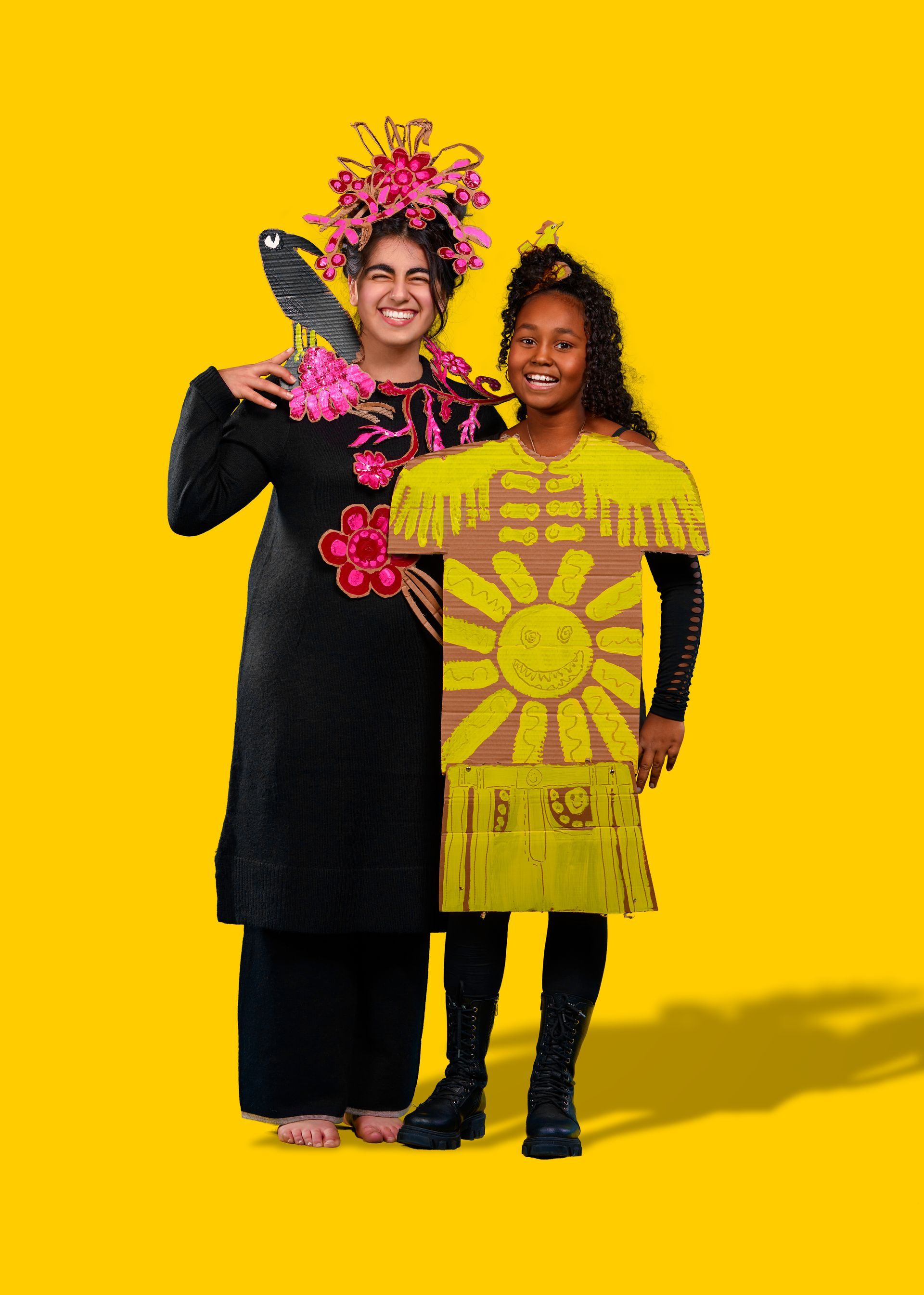 Twee meisjes in kostuum. Een draagt ​​een zwarte outfit met bloemenversieringen. De ander draagt ​​een kartonnen zonnepak.