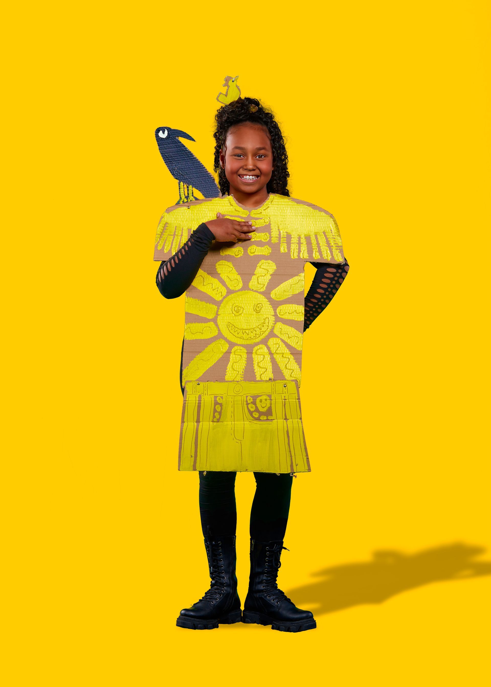 Jongere in een handgemaakt zonnepak van karton, geel en zwart, met een papieren raaf op de schouder.