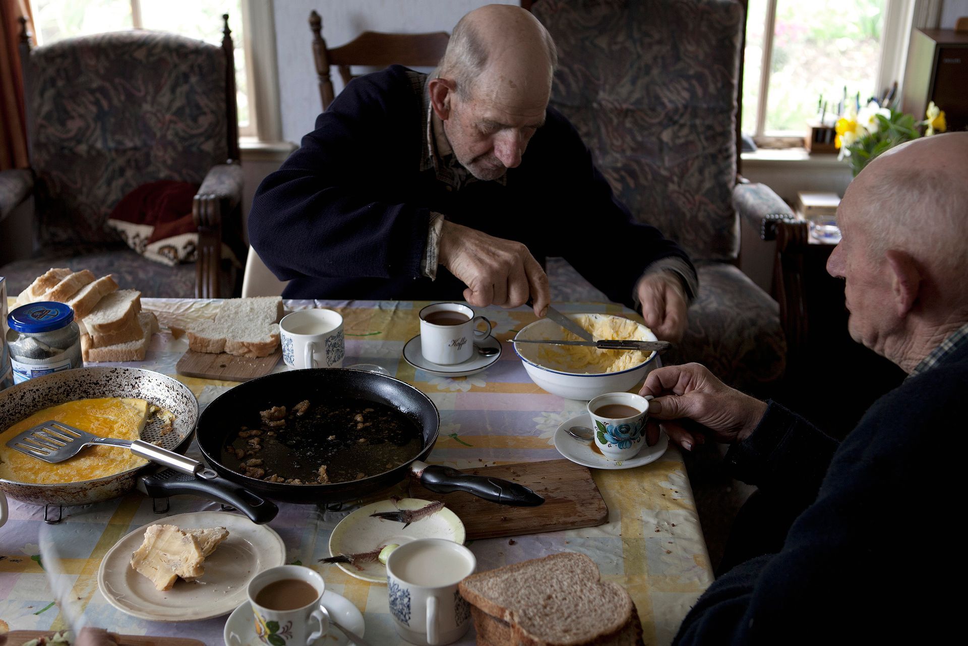 Twee broers aan een tafel; de een smeert het brood, de ander houdt een kop koffie vast .Foto Elise Klinkert