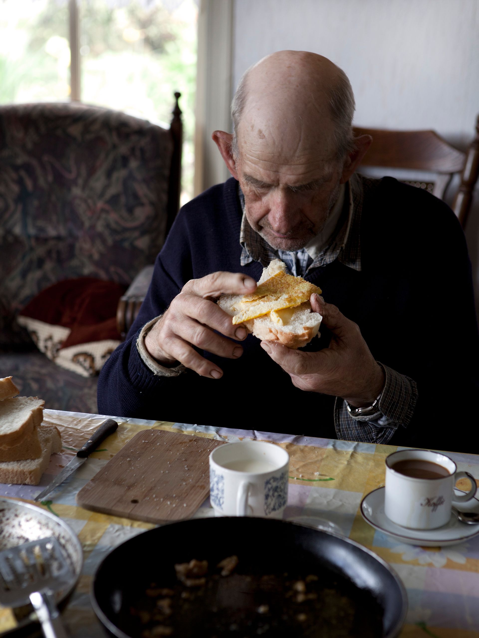 Een oudere man eet een boterham met ei aan een tafel. 