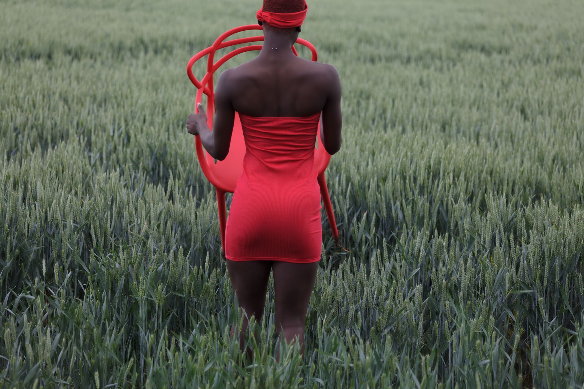 Vrouw in een rode jurk  draagt ​​een rode stoel door een groen veld. Foto Elise Klinkert