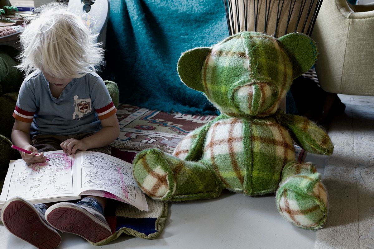 Een kind met blond haar kleurt in een kleurboek,  naast een geruite groene teddybeer, binnenshuis.