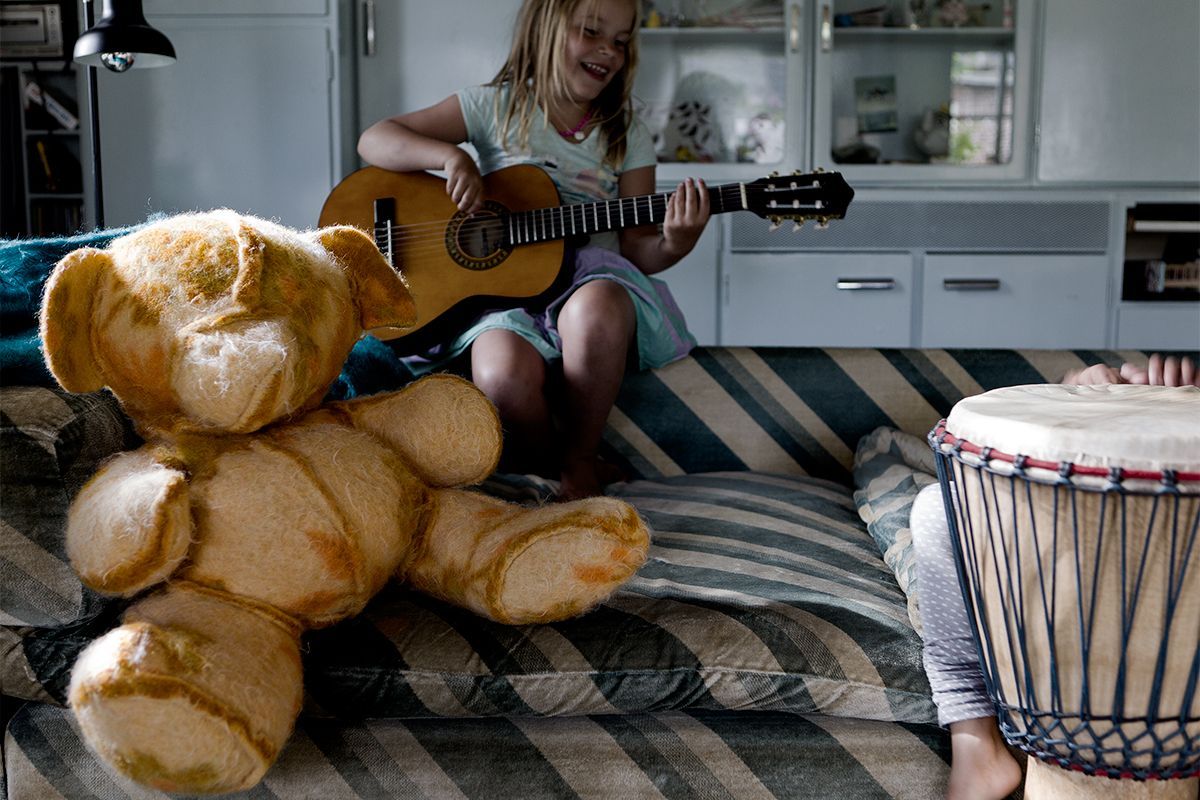 Meisje lacht terwijl ze gitaar speelt, teddybeer op gestreepte bank, trommel op de voorgrond.