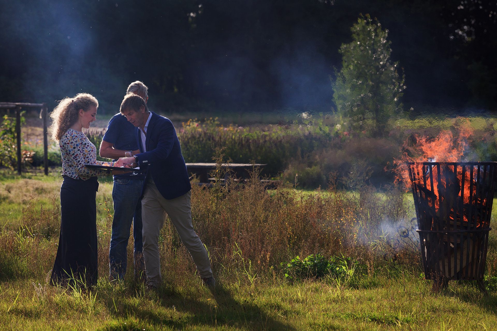 Drie mensen staan ​​bij een brandende vuurkorf in een tuin.