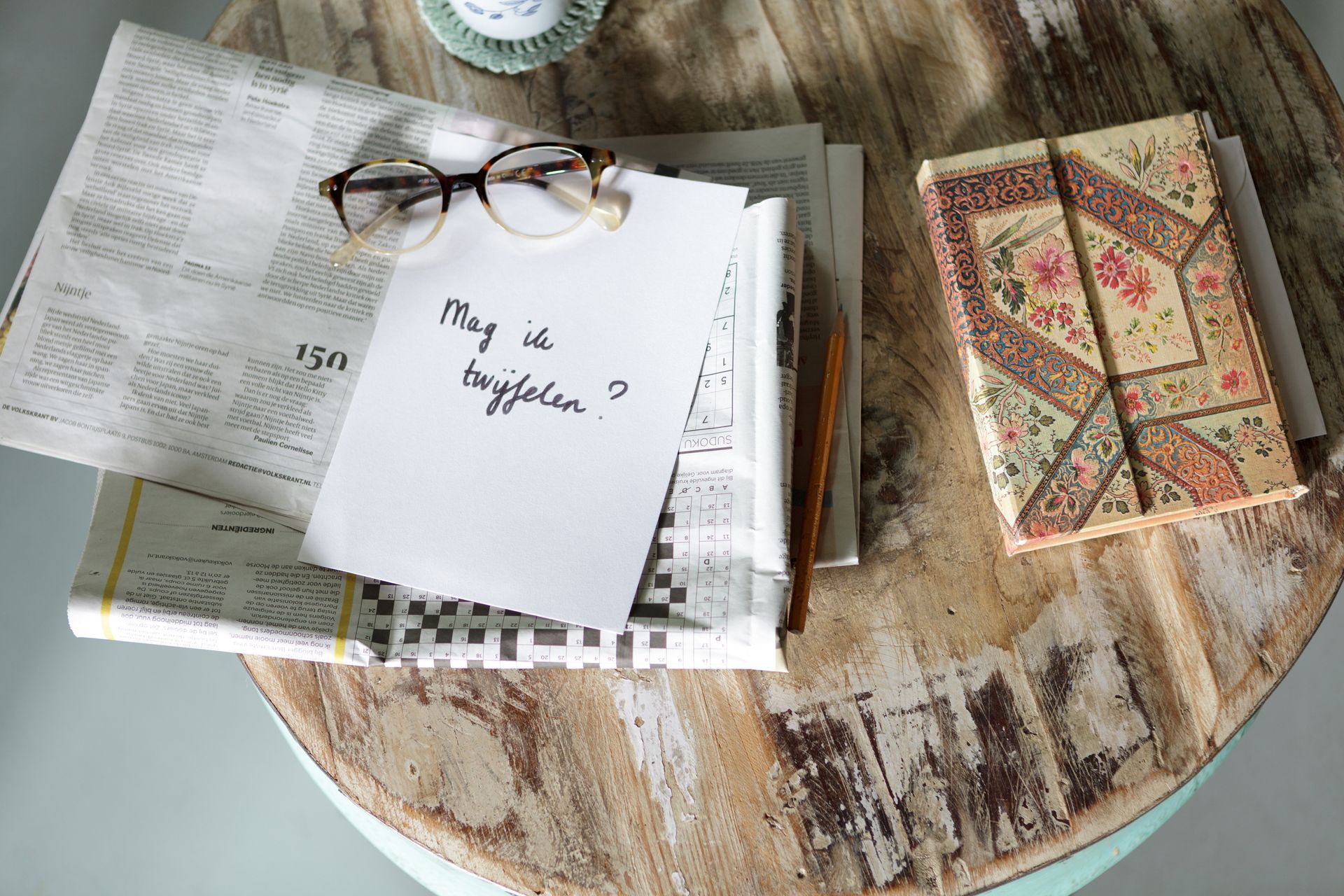 Een bril op een notitieblok naast een krant en een boek op een rustieke tafel. Foto Elise Klinkert