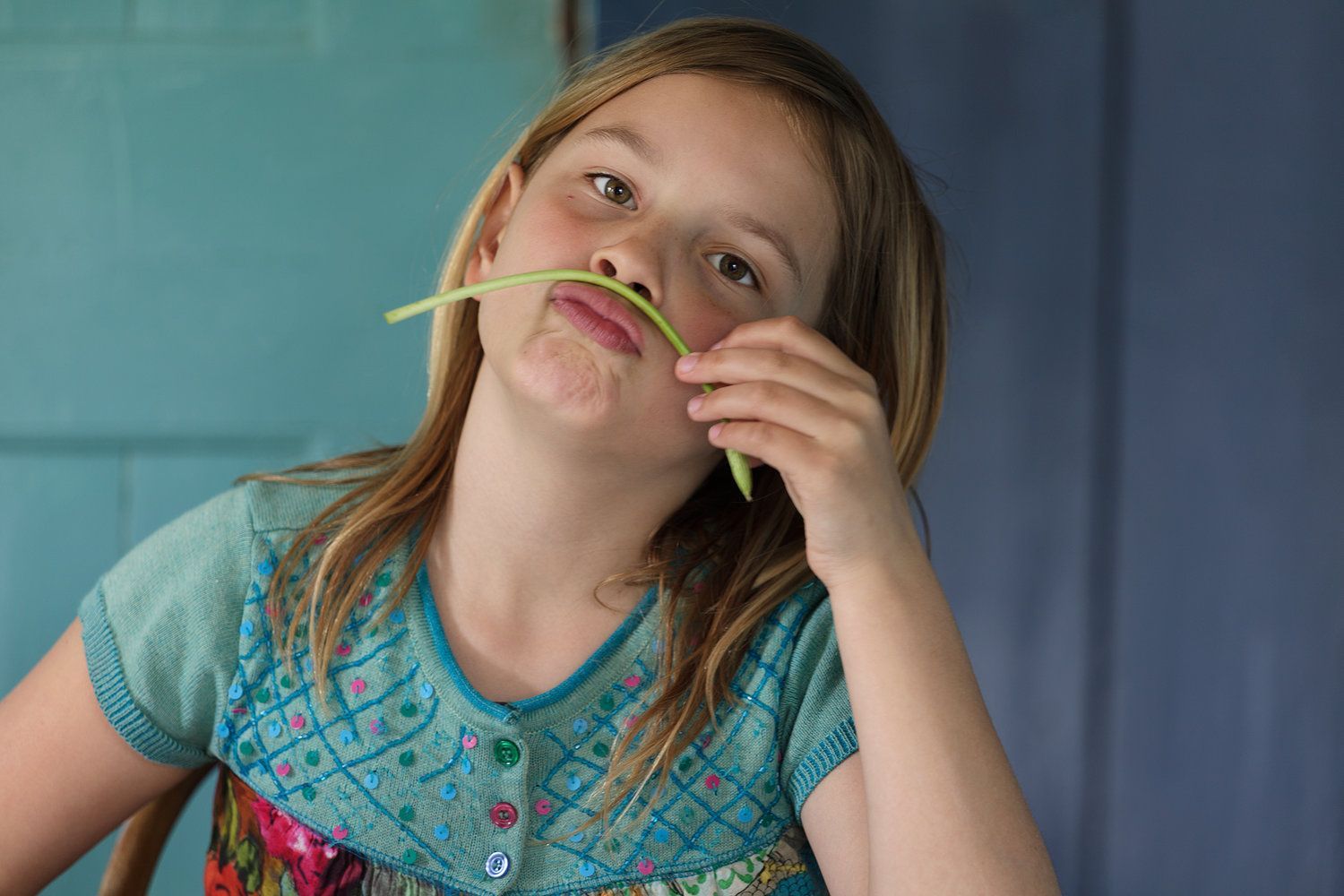 Meisje met lang blond haar maakt een snor met een groene asperge . Blauwe achtergrond. Foto Elise Klinkert