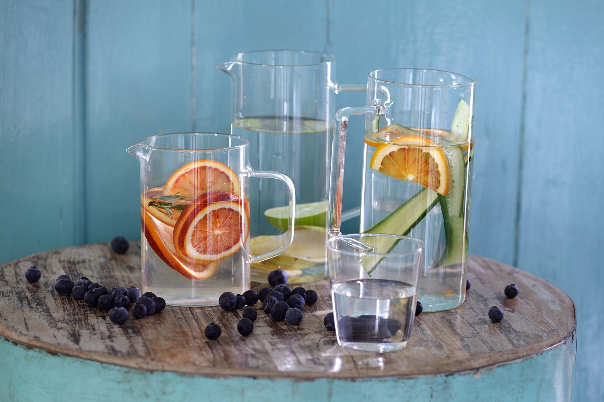 Waterkannen met fruitschijfjes en bosbessen op een houten tafel tegen een blauwe muur.