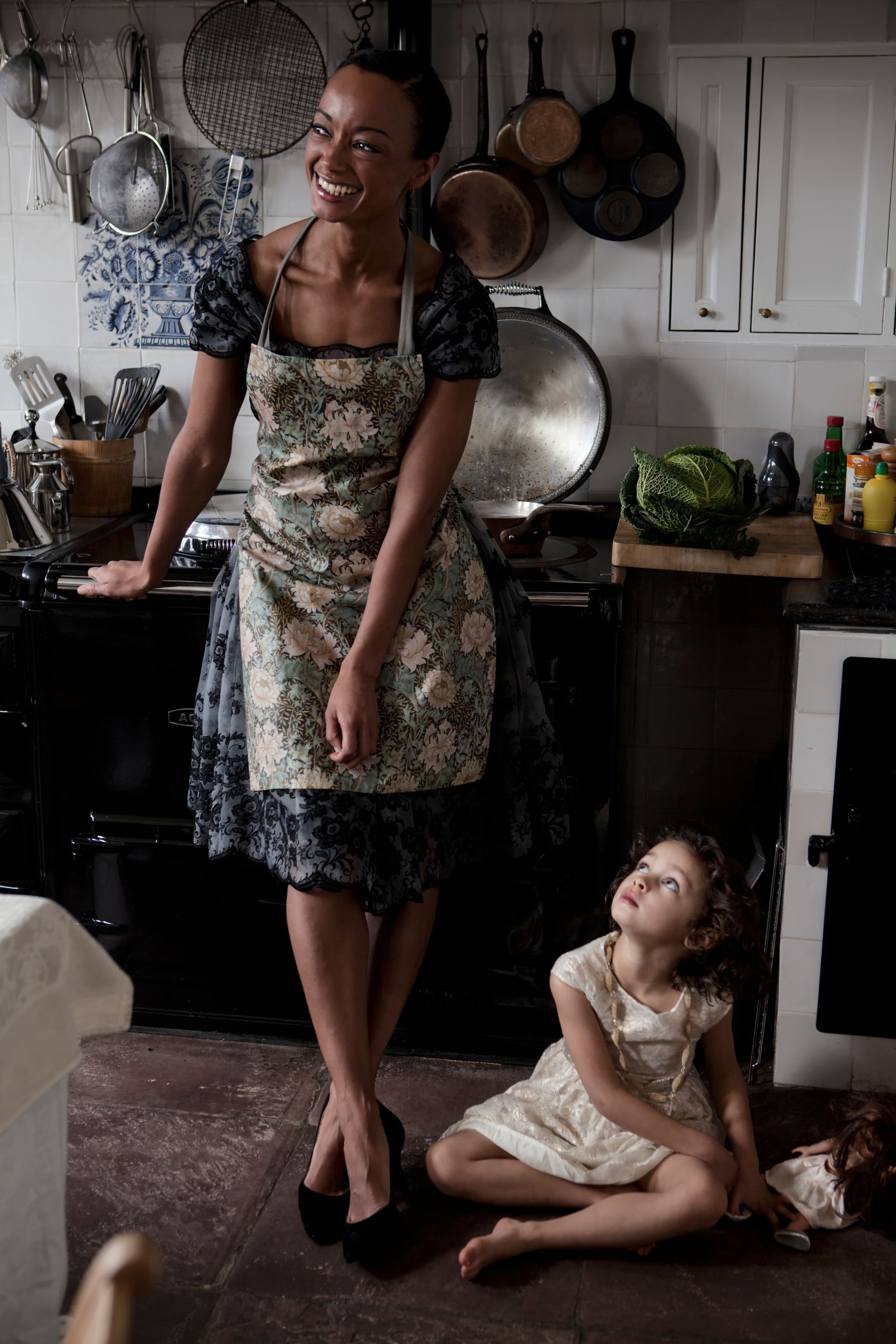 Vrouw met schort lacht in de keuken, kind kijkt omhoog. Potten hangen, kool op het aanrecht.Foto Elise Klinkert