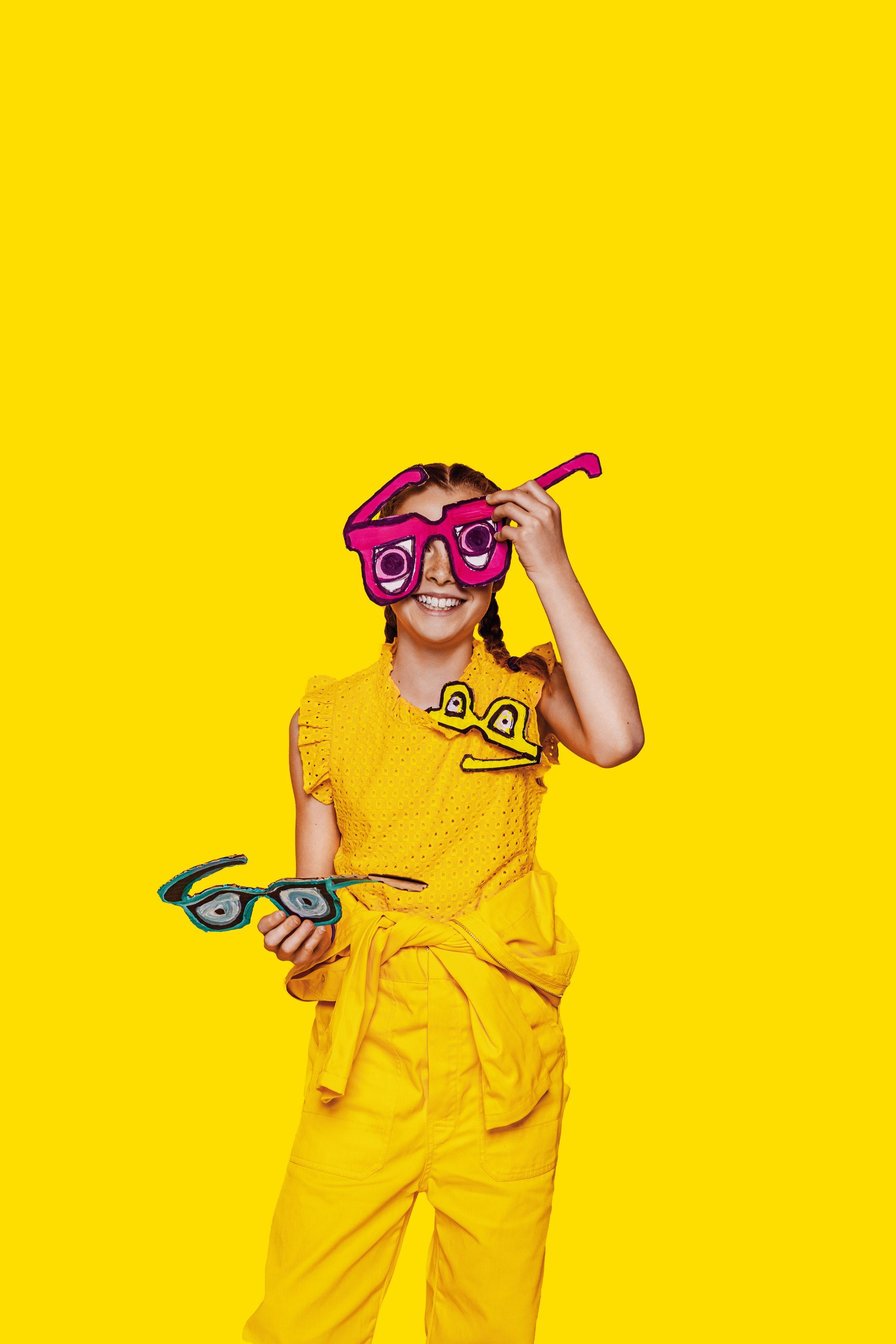 Meisje in een gele outfit met een grote roze bril, die een speelgoedbril vasthoudt op een gele achtergrond.