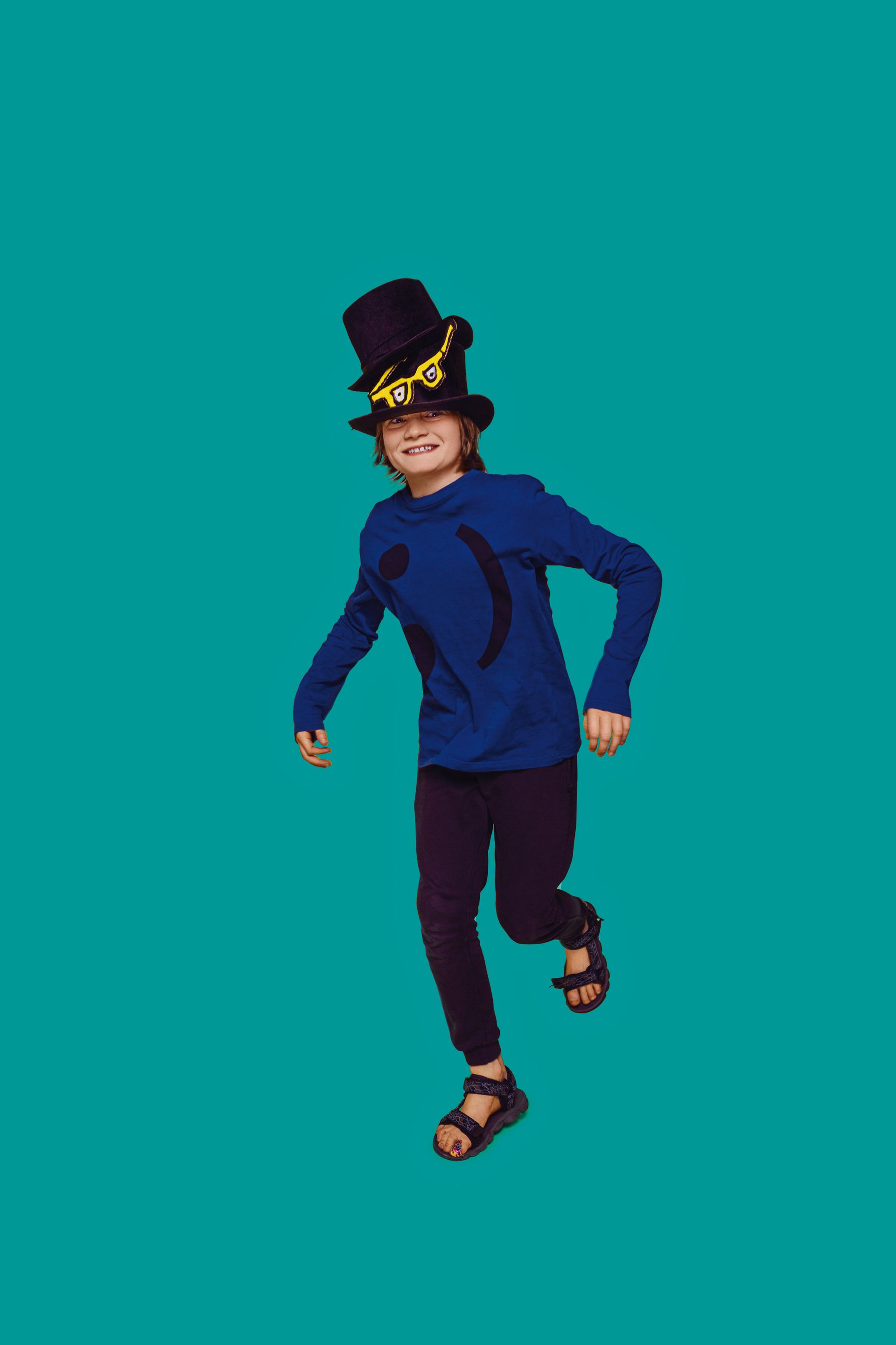 Een kind met een hoge hoed en een blauw shirt rent tegen een turquoise achtergrond.