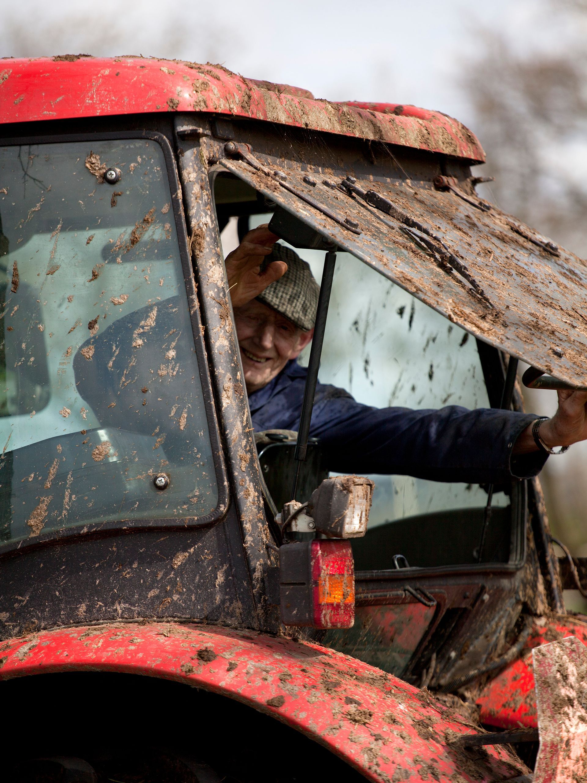 Een boer in een met modder besmeurde rode tractor kijkt met een glimlach uit het raam.