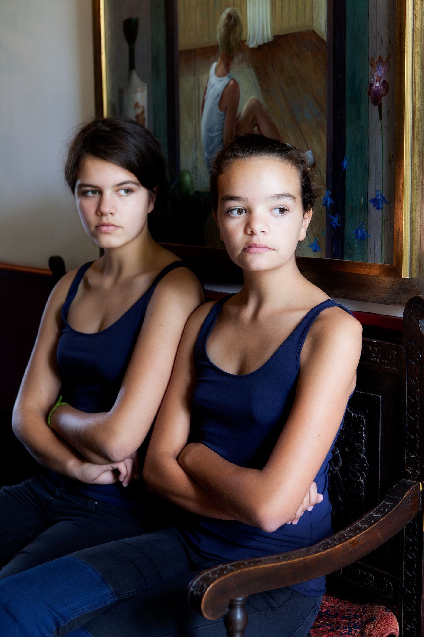 Twee tienermeisjes, armen over elkaar, in blauwe tanktops. Binnen, sierlijke meubels, schilderij op de achtergrond.