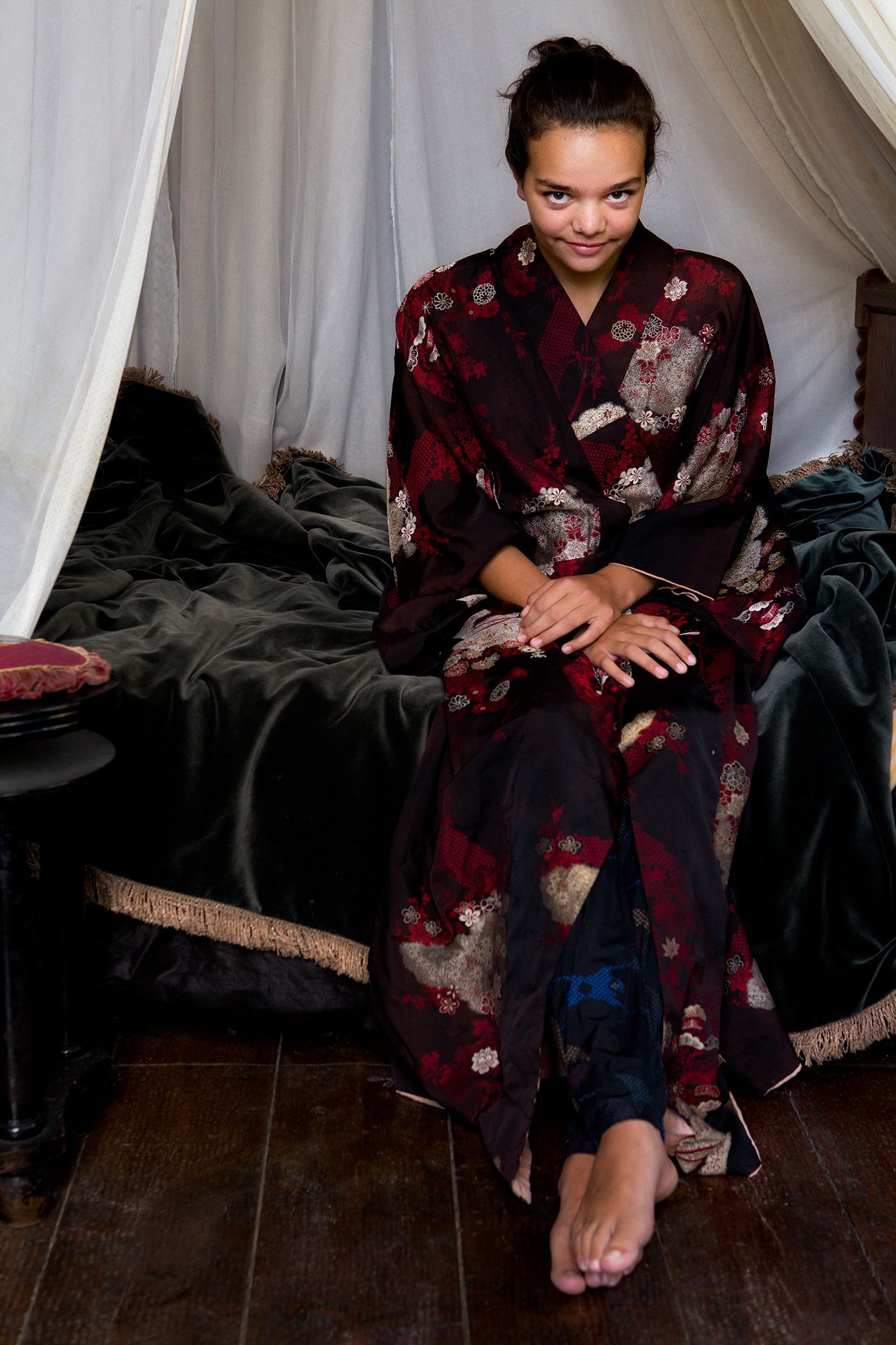 Een vrouw in kimono zit glimlachend op bed. Foto Elise Klinkert