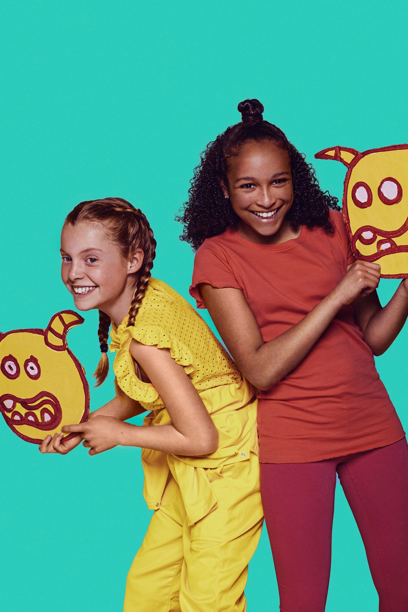 Twee lachende meisjes met gele monsterfiguurtjes in hun handen. Een in het geel, de ander in het rood. Turquoise achtergrond.