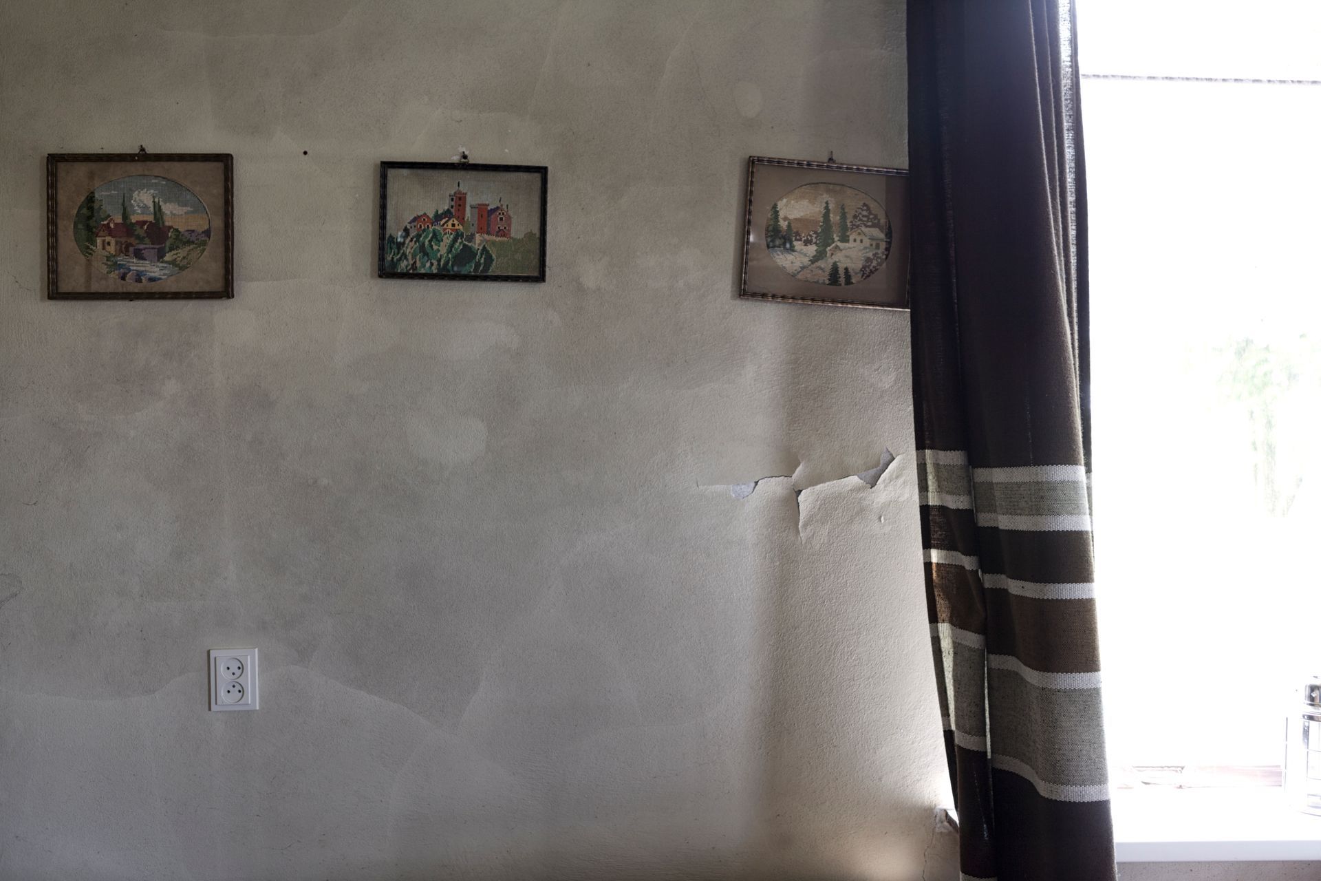 Drie ingelijste schilderijen aan een getextureerde muur. Een bruin gordijn bedekt de rechterkant. Er is een raam zichtbaar.
