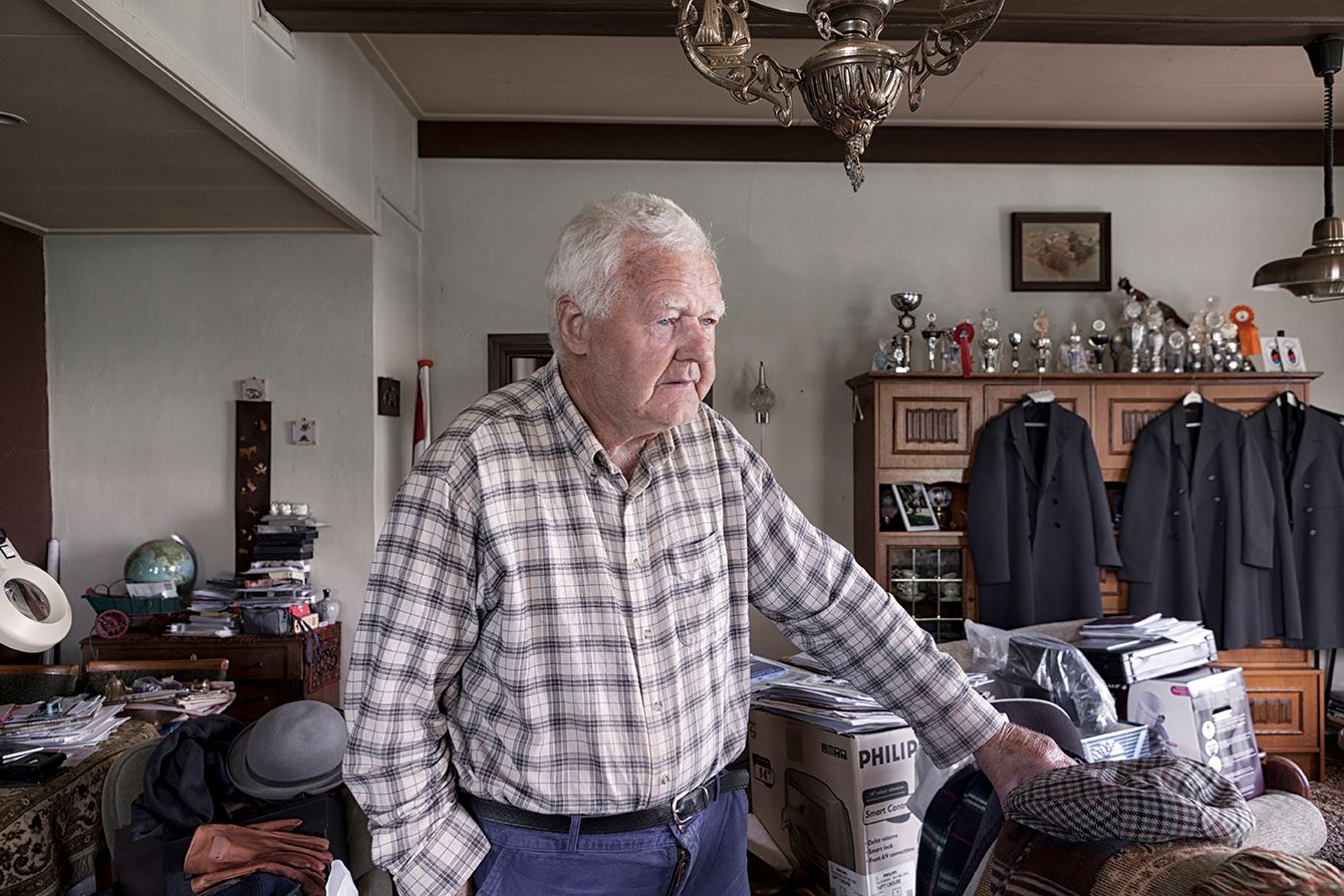 Een oudere man in een geruit overhemd in een rommelige kamer