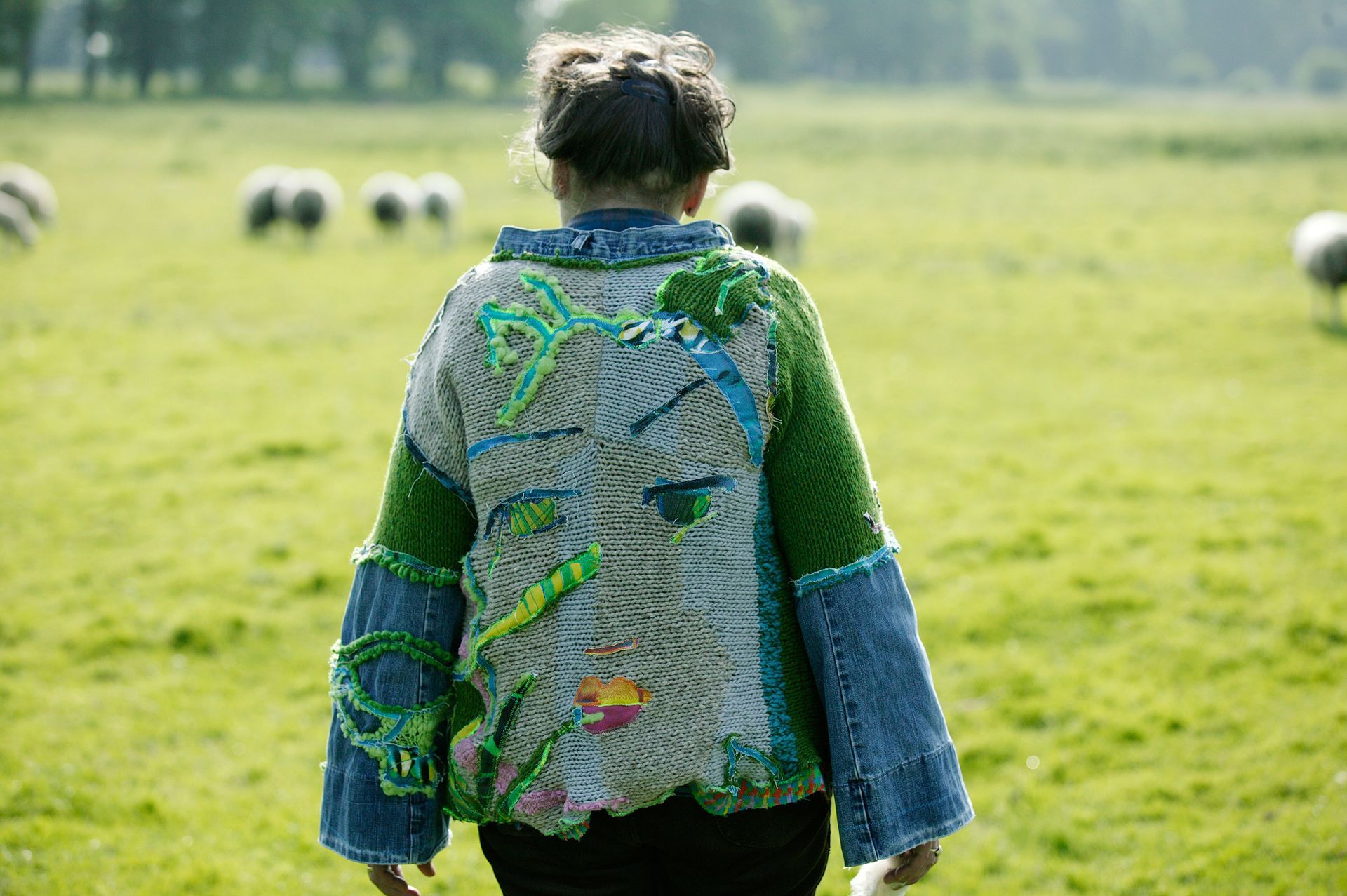 Een vrouw in een kleurrijke trui loopt door een veld, op de achtergrond grazen schapen.