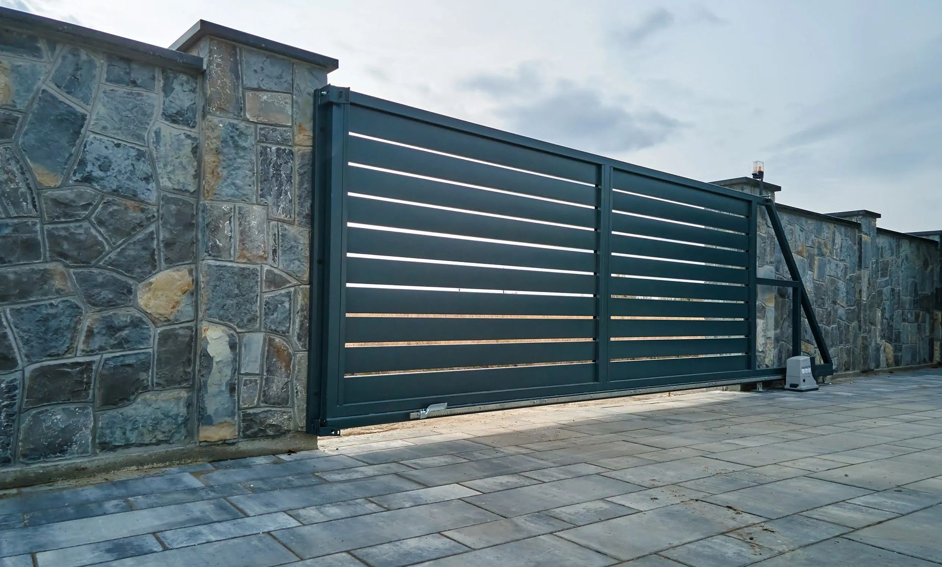 Dark green metal gate set in a stone wall beside a paved driveway under cloudy sky