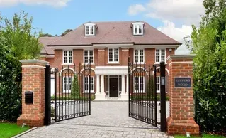 Large brick house with a gated front entrance and driveway, framed by trees and blue sky