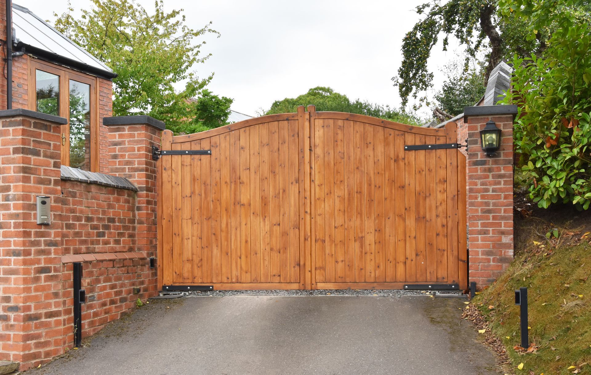 Wooden driveway gate between brick pillars beside a paved road and greenery