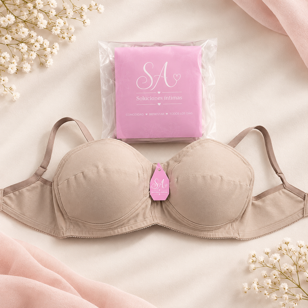 Sujetador beige con envoltorio rosa sobre un fondo de tela color crema suave, rodeado de flores blancas