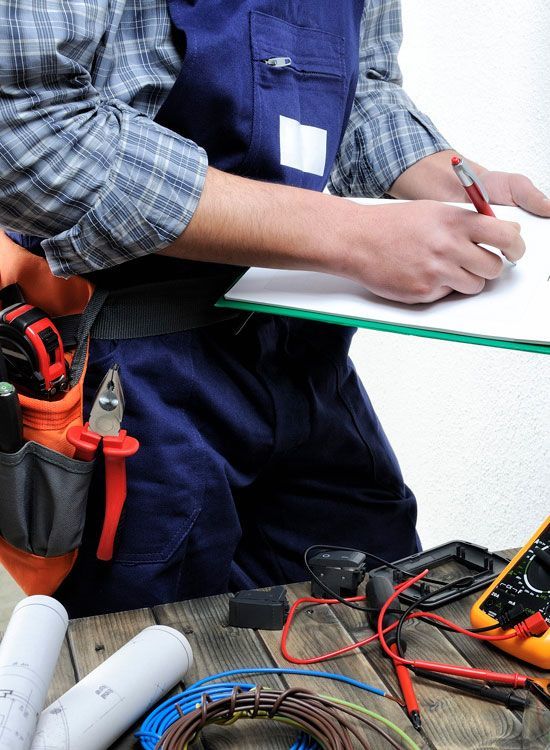 Electrician in blue overalls, writing on a clipboard at a workbench with tools and wires.
