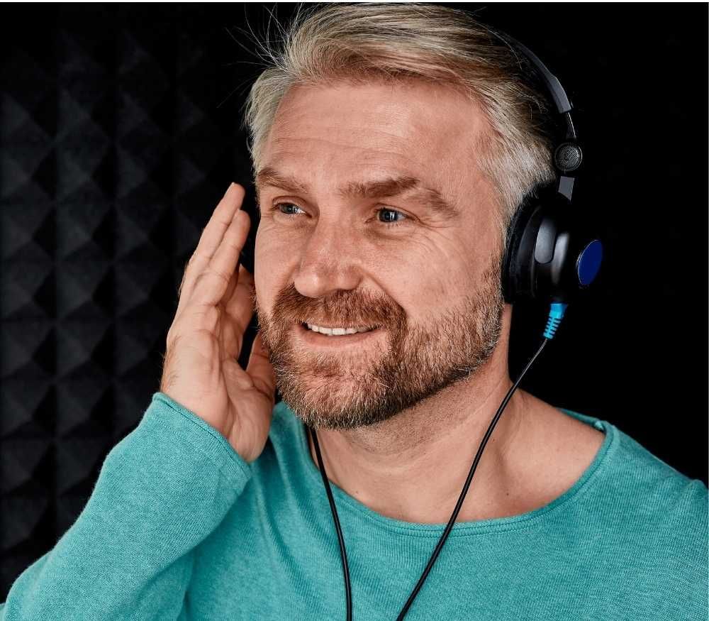Smiling man wearing headphones and a turquoise shirt, touching his face in a soundproof studio.