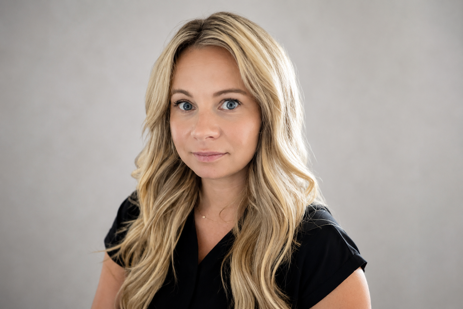 Portrait of a blonde person in a black top, smiling against a gray background