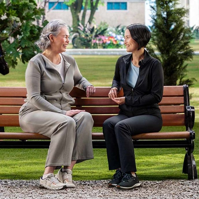 Two people talking on a park bench, smiling in a green garden setting.