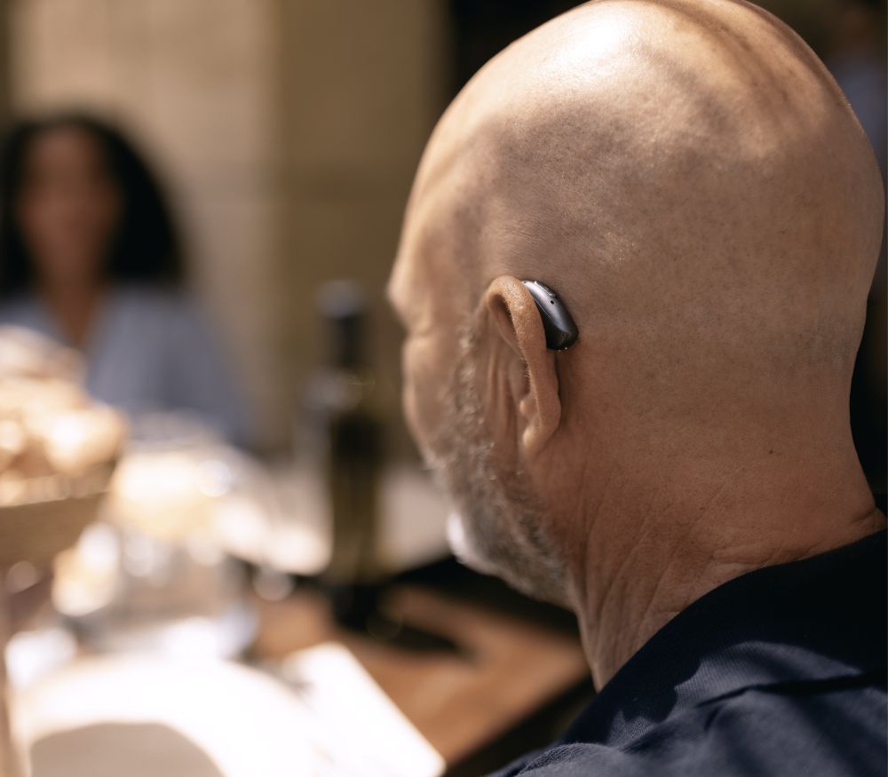Bald person wearing a hearing aid, seen from behind at a dining table