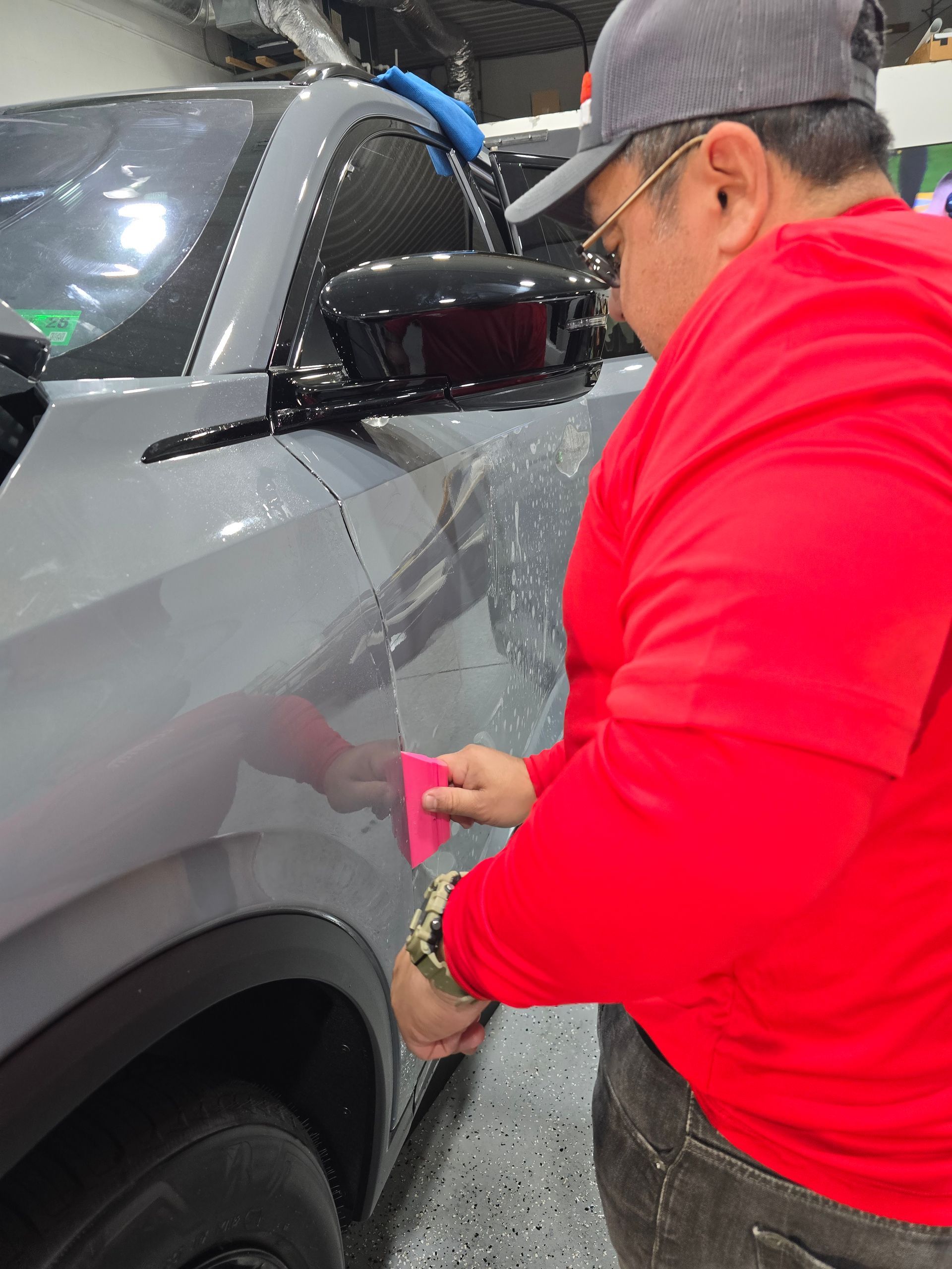 Person in red shirt polishing a gray car door with a pink cloth in a garage.