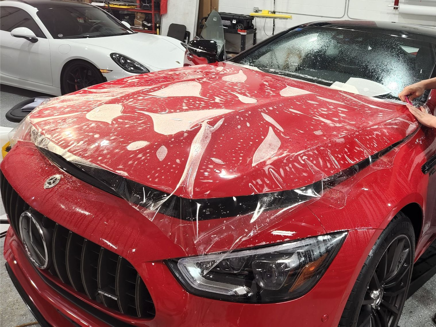 A man is covering the hood of a red car with plastic wrap.