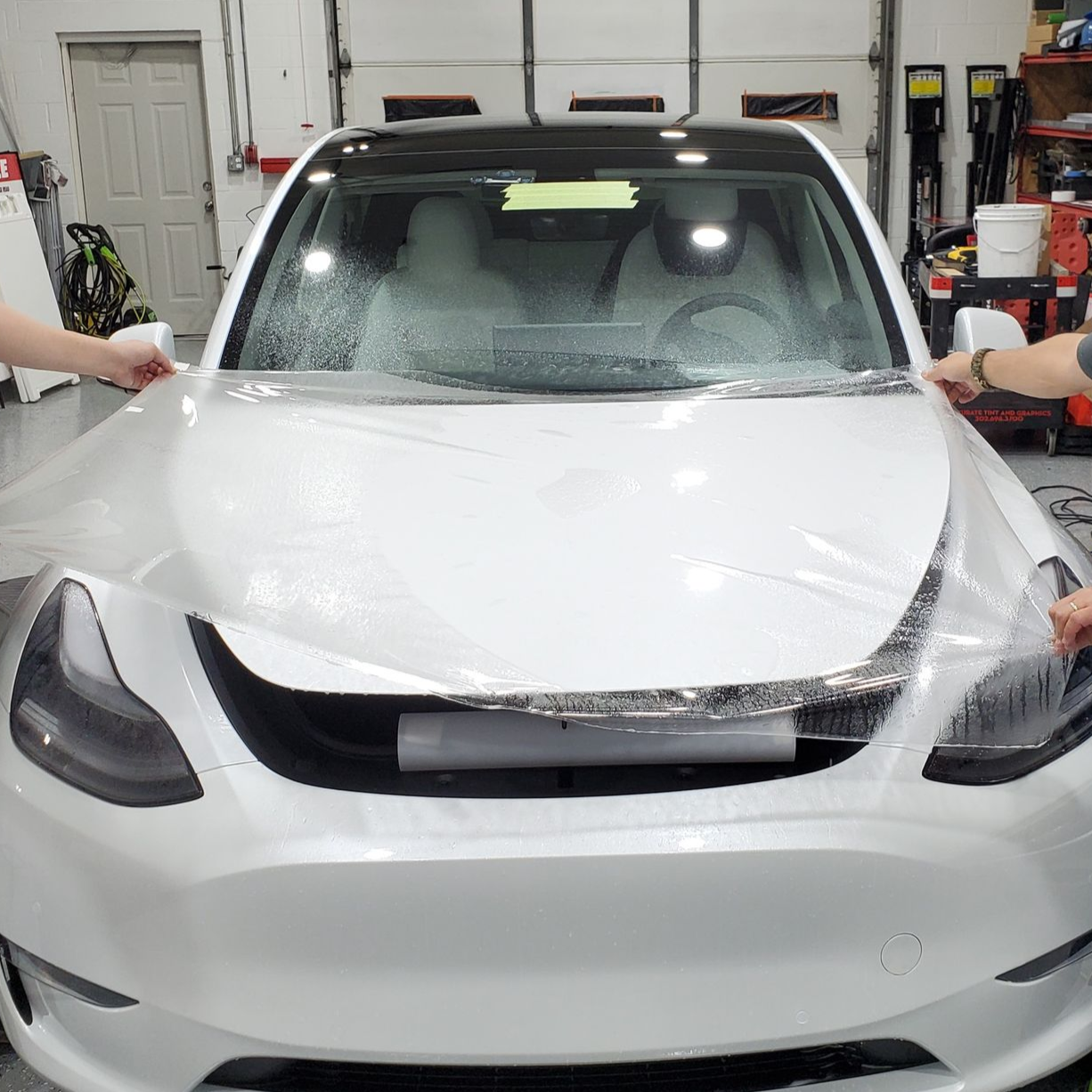 White car being wrapped in a garage, with two people holding the vinyl on either side.
