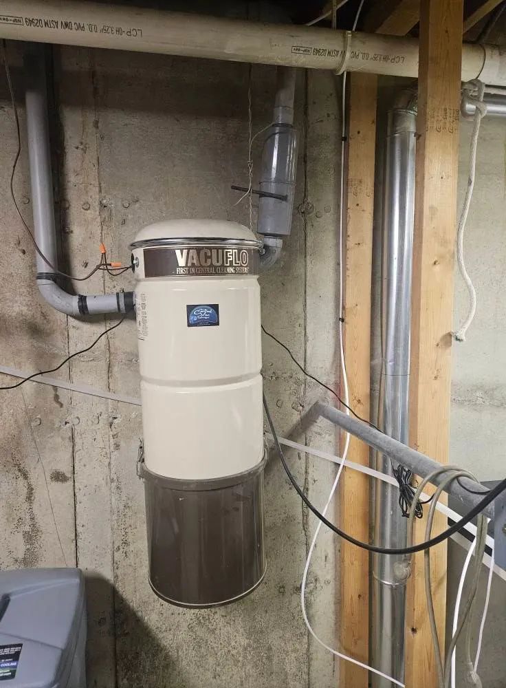 White cylindrical water heater in a basement beside exposed pipes and wooden framing.