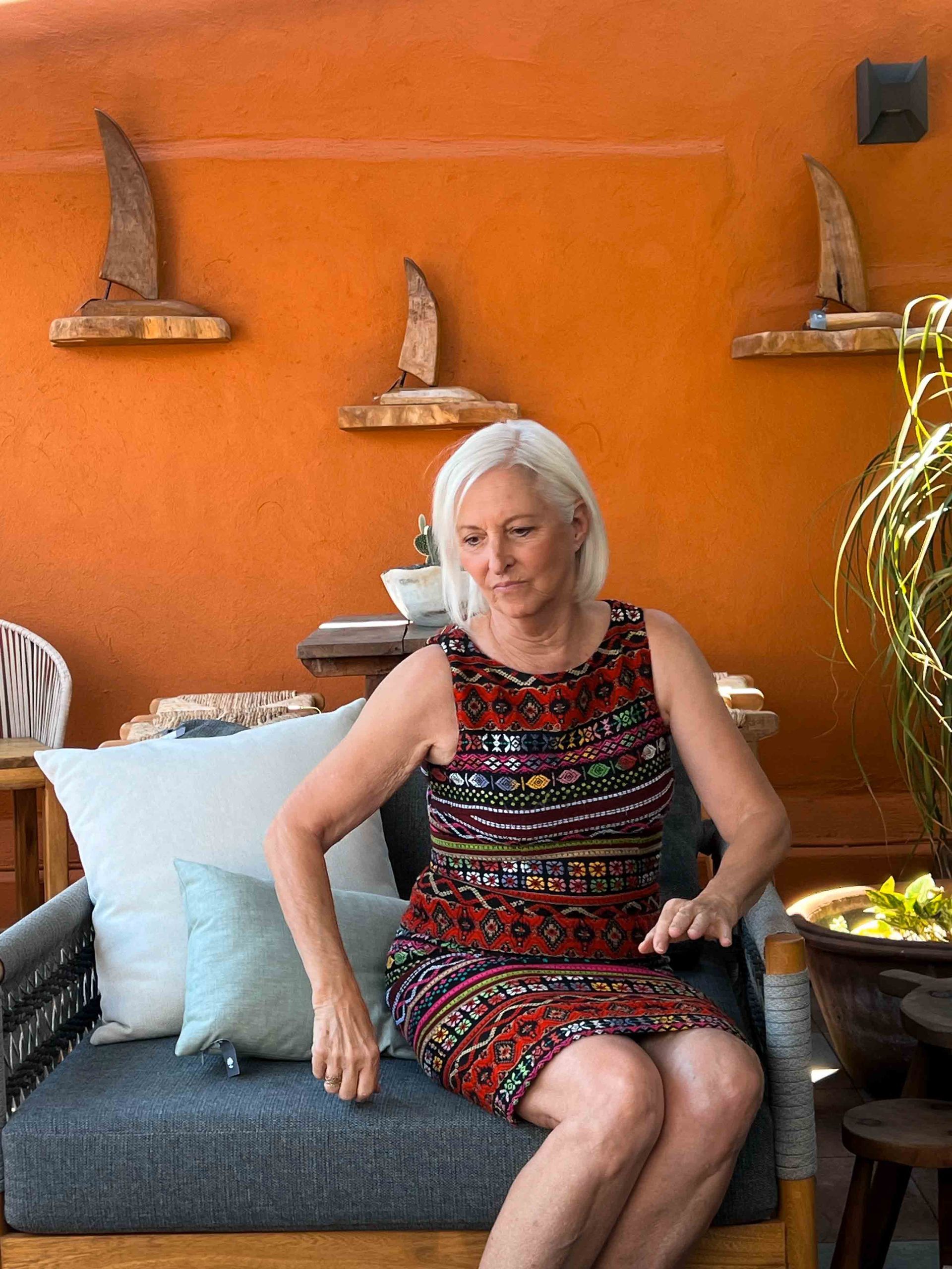 A woman in a dress is sitting in a chair in front of an orange wall.