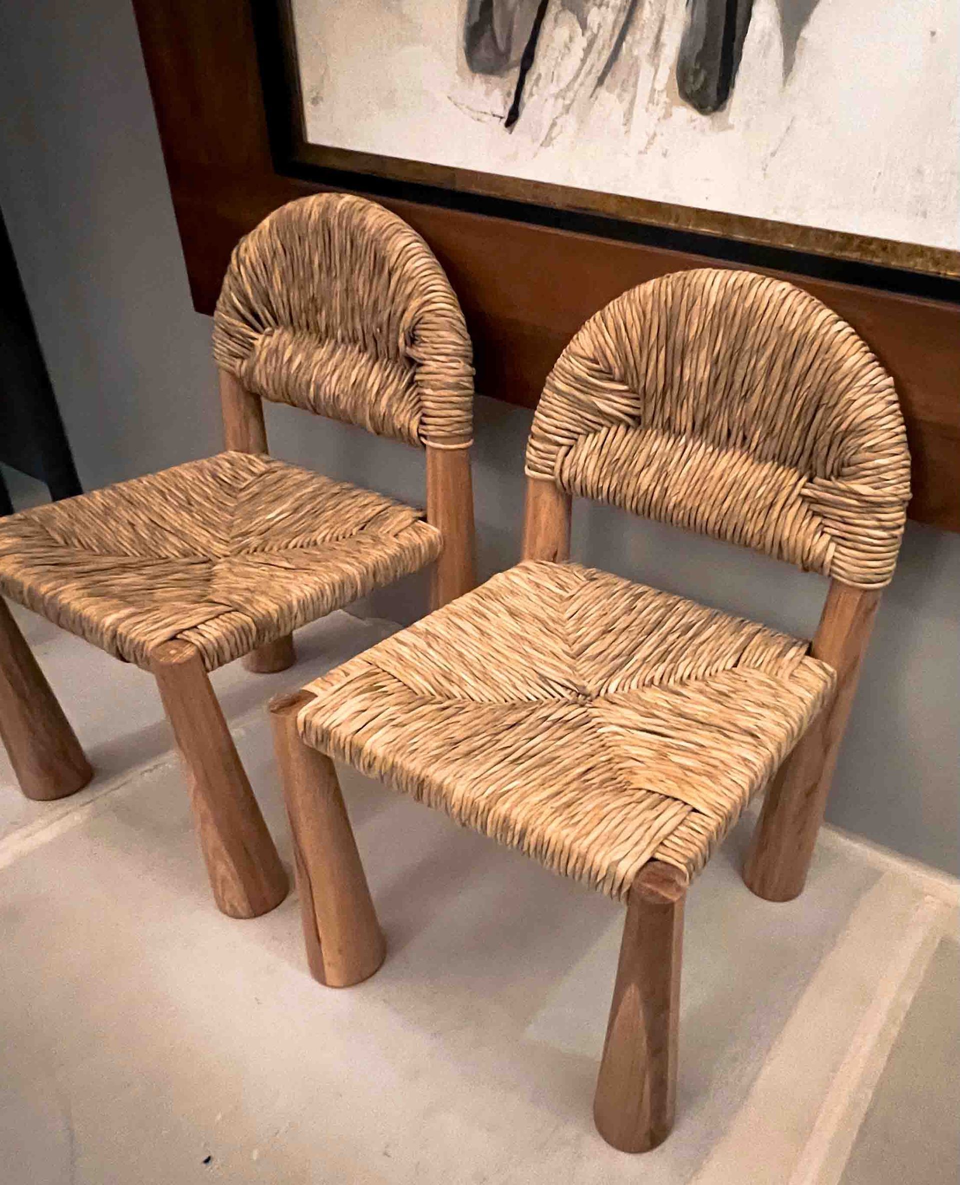 Two chairs made of straw and wood are sitting next to each other in front of a painting.