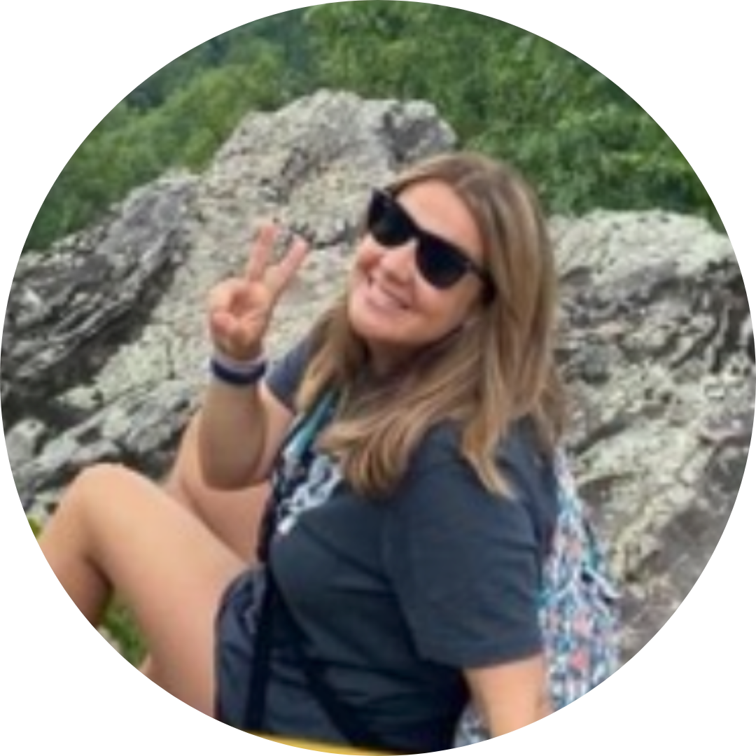 A smiling person wearing sunglasses makes a peace sign while sitting on a rocky outdoor trail.