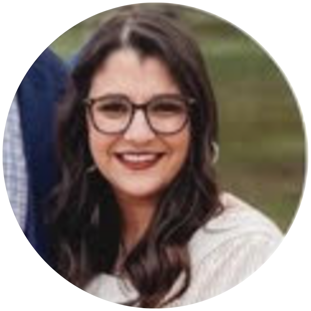 A smiling person with long, dark wavy hair and dark-rimmed glasses, wearing a light-colored top.