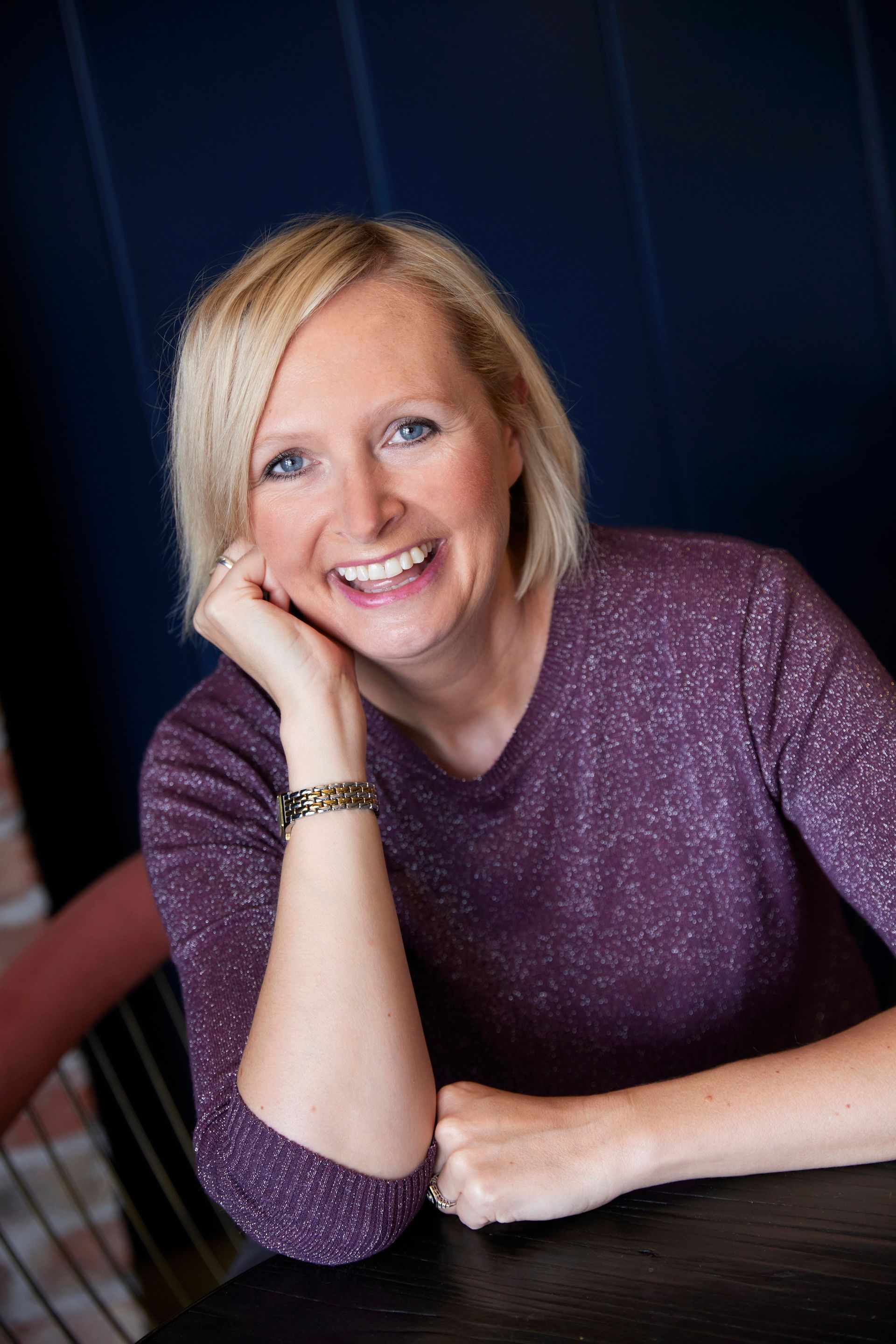 Woman with blonde hair smiles, resting her head on her hand at a dark table, wearing a purple sweater.
