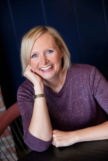 Woman with short blonde hair smiles, resting chin on hand at a table, wearing a purple sweater.