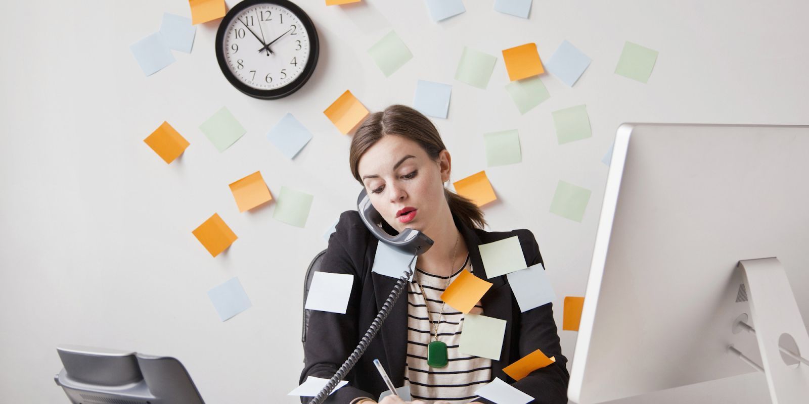 Overworked office worker on the phone at a desk, surrounded by sticky notes and a clock on the wall