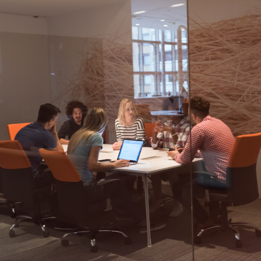 People seated around a table in a modern office, engaged in a discussion. Laptop, orange chairs.