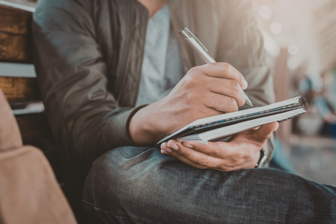 Person writing in a notebook while seated, wearing a dark jacket and jeans, with a soft blurred background