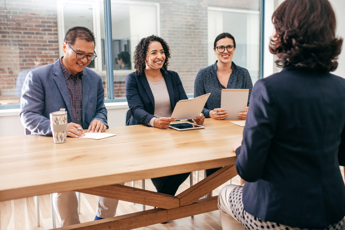 Group meeting in a bright office, with three people seated at a table facing a presenter.