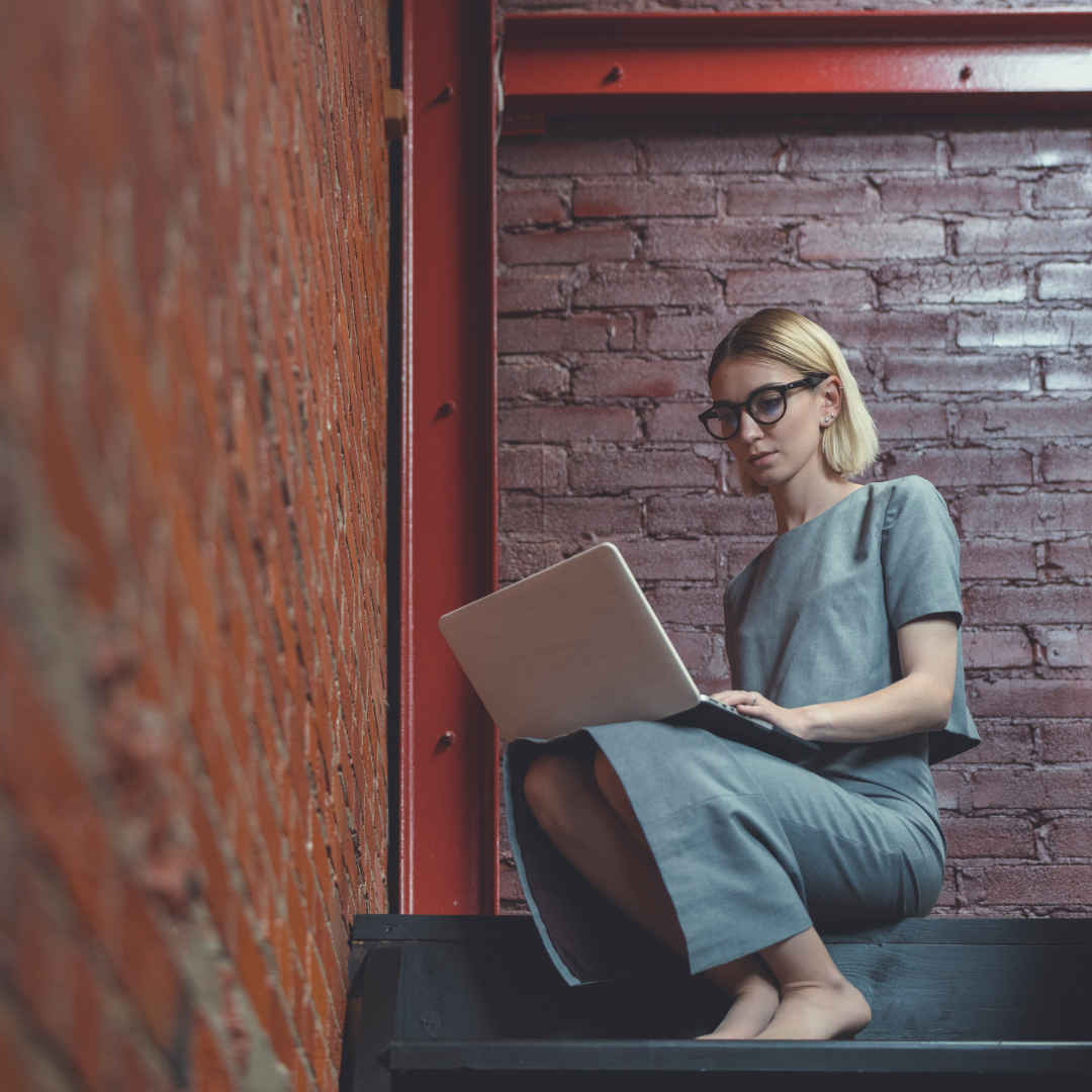 Woman with glasses using laptop, sitting on steps near a brick wall, wearing gray dress.