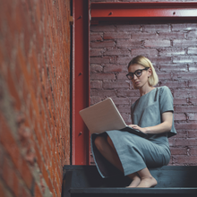Blonde woman with glasses, barefoot, sitting on steps, working on a laptop against a brick wall.