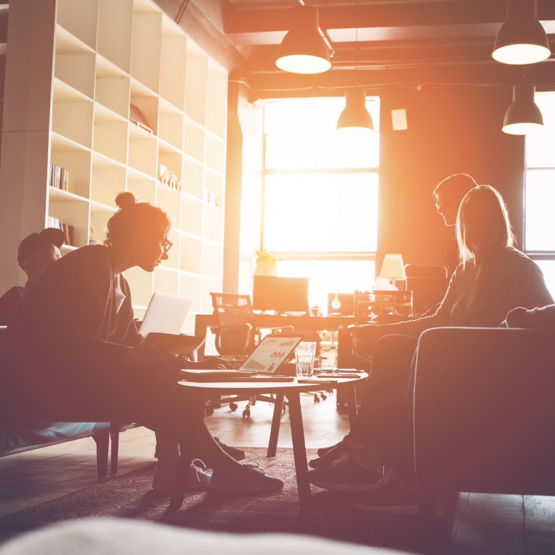 People working collaboratively in a bright office; one person at a laptop, others seated and conversing.