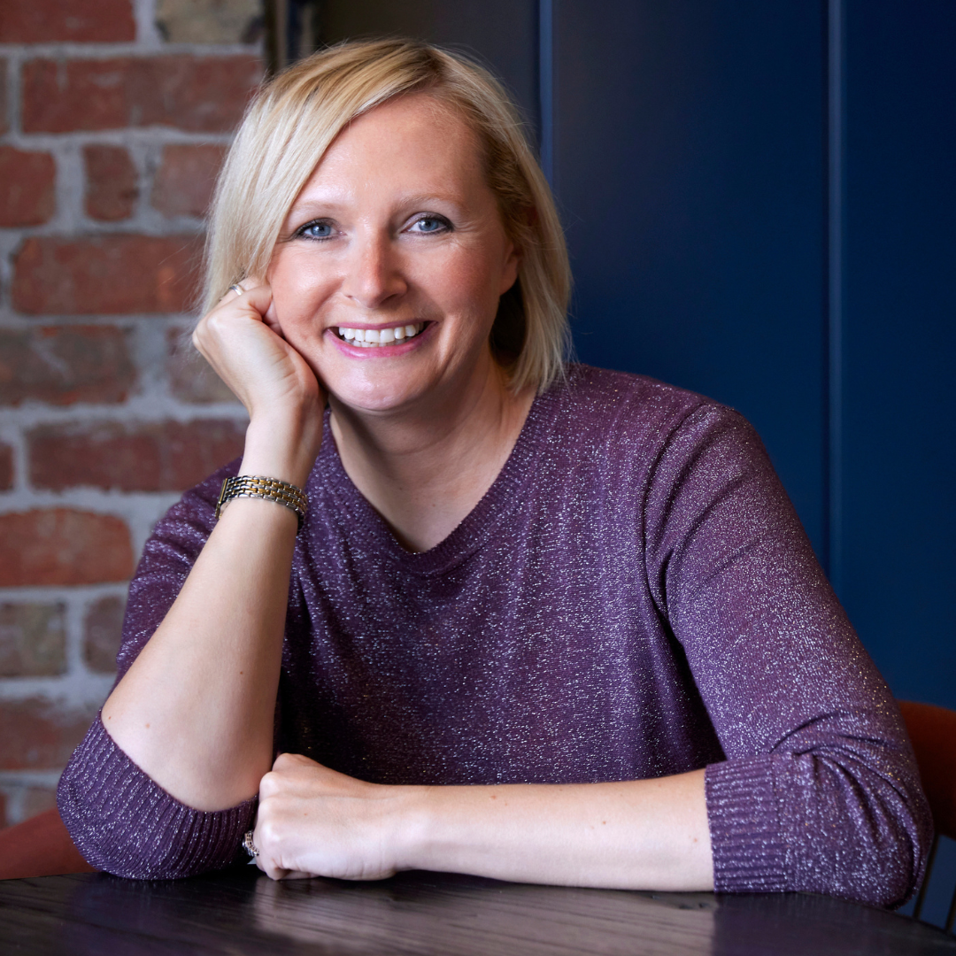 Woman with blonde hair smiles, resting chin on hand at a table, wearing a purple sweater, in front of a brick wall.