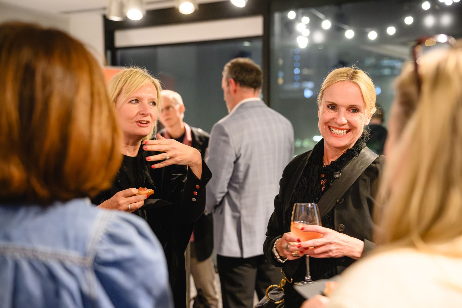 Guests chatting at a warmly lit cocktail party, with one woman holding a drink and smiling at the camera.
